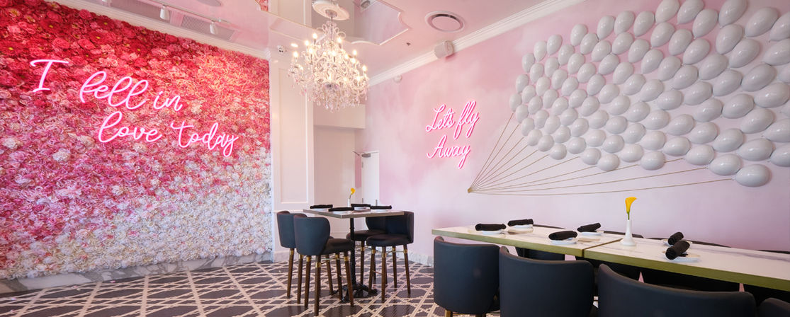 Playful pink cafe interior with a gradient floral wall and neon sign reading I fell in love today, balloon wall art with neon Let's fly away, chandelier, teal dining chairs and set tables