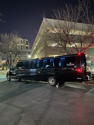 Black stretch party bus with blue interior lights parked near a multi-level parking garage and bare trees on a city street at night.