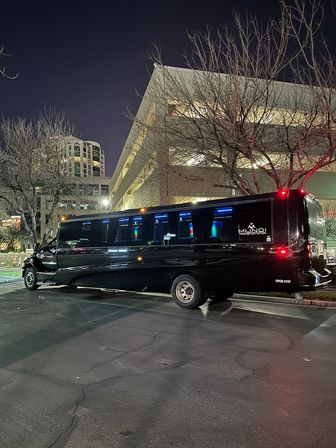 Black stretch party bus with blue interior lights parked near a multi-level parking garage and bare trees on a city street at night.