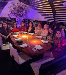 Group of women enjoying a night out at an upscale restaurant lounge with purple mood lighting, seated around a round wooden table set with menus, glasses and a large pink floral centerpiece.