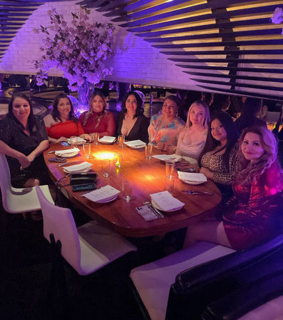 Group of women enjoying a night out at an upscale restaurant lounge with purple mood lighting, seated around a round wooden table set with menus, glasses and a large pink floral centerpiece.