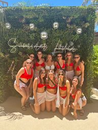 Outdoor pool-party photo: group of women in matching red bikinis and white sarongs posing in front of a lush green photo wall with a neon "Summer Lovin'" sign and hanging disco balls on a sunny day.