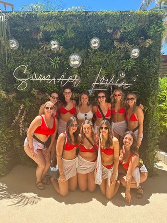 Outdoor pool-party photo: group of women in matching red bikinis and white sarongs posing in front of a lush green photo wall with a neon "Summer Lovin'" sign and hanging disco balls on a sunny day.
