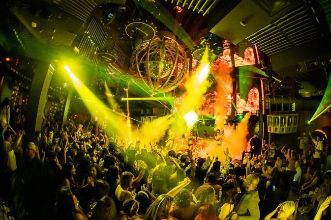 Packed nightclub dance floor with DJ booth, disco orb overhead, green-yellow spotlights, lasers and smoke creating a high-energy party atmosphere