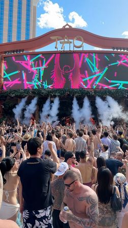 Sunlit Las Vegas pool dayclub party with a packed crowd of swimsuit-clad and shirtless guests raising hands in front of a large LED DJ stage showing neon visuals and plumes of smoke