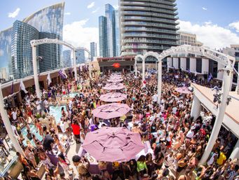 Lively rooftop pool party with hundreds in swimwear, rows of purple umbrellas, pools and a downtown high-rise skyline.