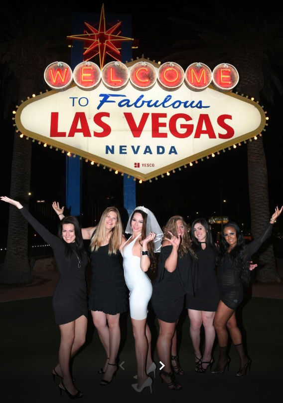 Six women in evening dresses, one wearing a white dress and veil, posing and cheering beneath the illuminated 'Welcome to Fabulous Las Vegas Nevada' sign at night.