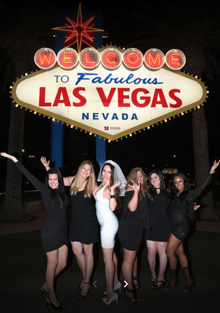 Six women in evening dresses, one wearing a white dress and veil, posing and cheering beneath the illuminated 'Welcome to Fabulous Las Vegas Nevada' sign at night.