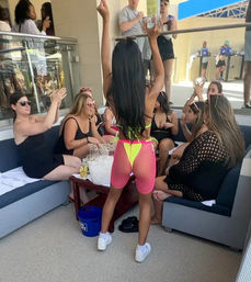 Pool club cabana party with friends cheering as a woman in neon pink fishnet shorts and yellow bikini raises her arms over a table of drinks and ice.