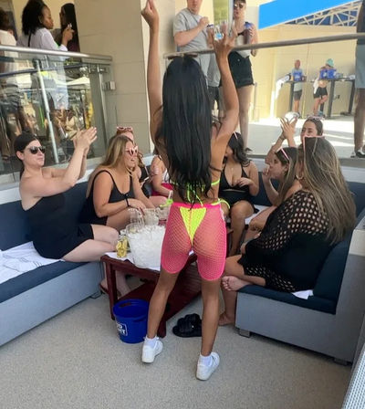 Pool club cabana party with friends cheering as a woman in neon pink fishnet shorts and yellow bikini raises her arms over a table of drinks and ice.