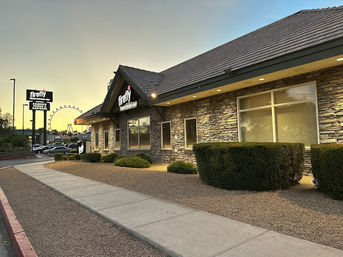 Dusk view of a one-story stone-faced roadside restaurant with warm exterior lights, gravel landscaping and trimmed bushes along a sidewalk, with a Ferris wheel glowing on the horizon.