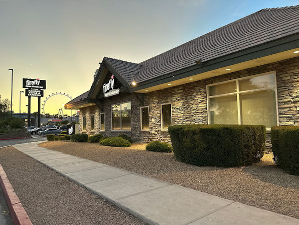 Sunset exterior of a single-story stone-faced restaurant with glowing sign, desert rock landscaping and trimmed shrubs along a sidewalk, parking lot and distant Ferris wheel in the entertainment-district background.