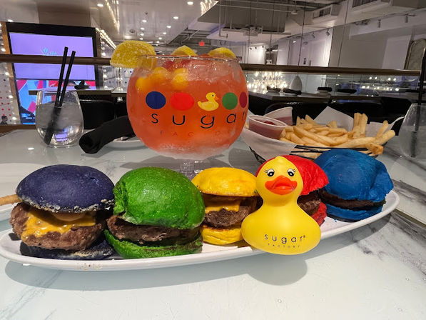 Plate of colorful mini cheeseburger sliders with purple, green, yellow, red and blue buns, fries and a yellow rubber duck, plus a large fruity punch bowl with lemon slices on a marble table in an indoor restaurant setting.