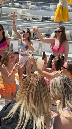 Sunny rooftop pool lounge with a group of women toasting shots, bachelorette in white sash and heart sunglasses celebrating with friends.