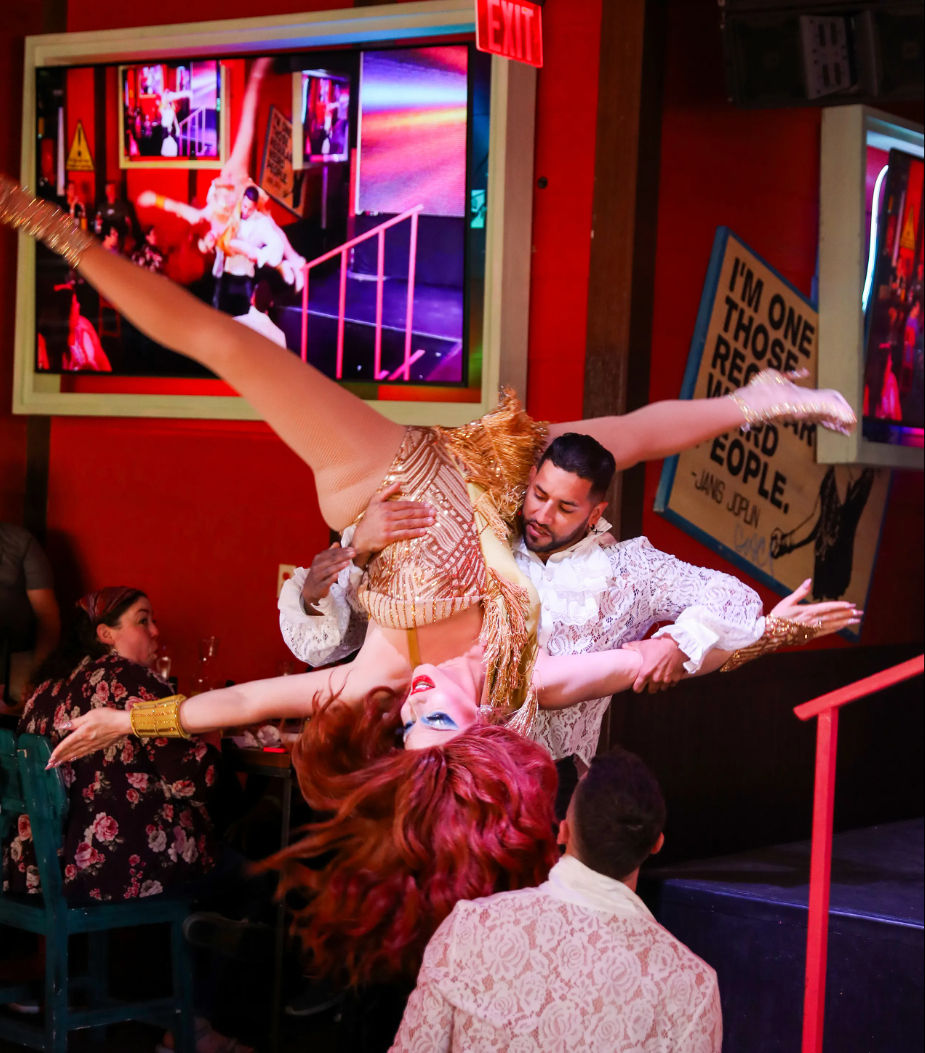 Red‑haired cabaret performer in a gold sequin costume flipped upside down by her partner in a white lace shirt during a lively nightclub/dinner show with colorful screens and seated audience.
