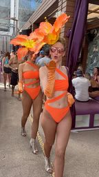 Two performers in bright orange cutout bikinis, fishnet tights and oversized orange floral headpieces walking and blowing a kiss at a lively outdoor pool party cabana.