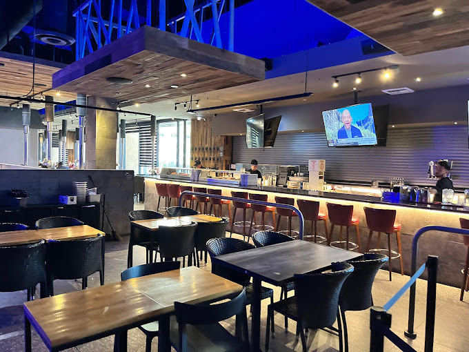 Modern urban bar and lounge interior with wooden tables and wicker chairs, long illuminated counter with red bar stools, blue accent ceiling lighting and wall-mounted TVs.