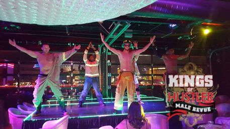 Neon-lit nightclub stage with shirtless male revue dancers in firefighter-themed pants striking poses as an audience watches, city lights glowing outside.