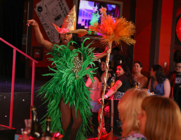 Drag performer in a green feather costume and gold wide-brim hat holding a sparkly orange feathered staff entertaining a seated audience at a lively nightclub cabaret