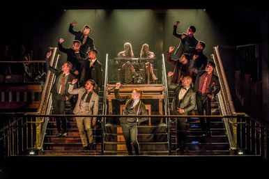 Theatre stage musical: a chorus of men in suits striking a synchronized pose on a grand dual staircase under warm spotlights, two women seated at the top.