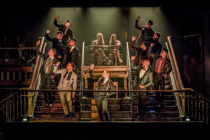 Theatre stage musical: a chorus of men in suits striking a synchronized pose on a grand dual staircase under warm spotlights, two women seated at the top.