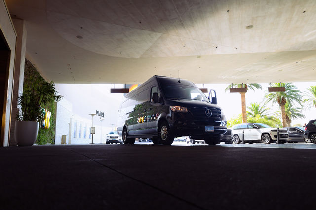 Black Mercedes Sprinter hotel shuttle parked under a porte-cochere at a sunny, palm‑lined resort entrance — valet area with other cars nearby.