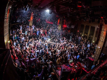 Packed nightclub dance floor with DJ booth, confetti falling and partygoers waving glow sticks under red and white lights — high-energy urban nightlife party scene.