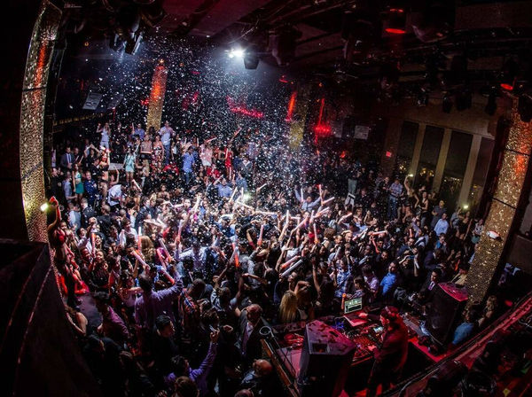 Packed nightclub dance floor with DJ booth, confetti falling and partygoers waving glow sticks under red and white lights — high-energy urban nightlife party scene.