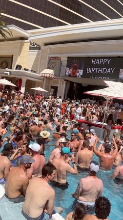Lively daytime resort pool party with dozens of people in swimwear packed into a shallow pool, umbrellas and cabanas, a DJ stage and a large LED screen displaying a birthday message.