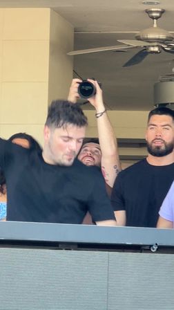 Group on a venue balcony — a man lifts a camera overhead to shoot while two men in black shirts stand nearby under a ceiling fan.