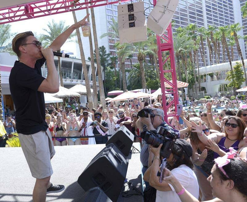 Daytime pool party concert at a sunny resort: performer on stage with a microphone and drink pumping up a cheering crowd of fans and photographers amid palm trees, umbrellas and hotel high-rises.