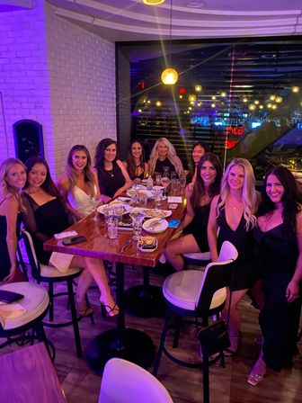 Eleven women in evening dresses smiling around a high wooden table with plates and cocktails, friends’ night out at a modern restaurant and bar with purple mood lighting and hanging globe lights reflecting in the window.