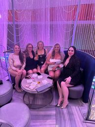 Five women in cocktail dresses at a bachelorette party in a purple-lit lounge with beaded curtain backdrop, smiling and holding cocktails around a small table.