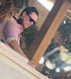 Outdoor DJ in patterned shirt with sunglasses and headphones leaning over a booth on a sunny terrace with wooden posts and greenery, poolside lounge vibe