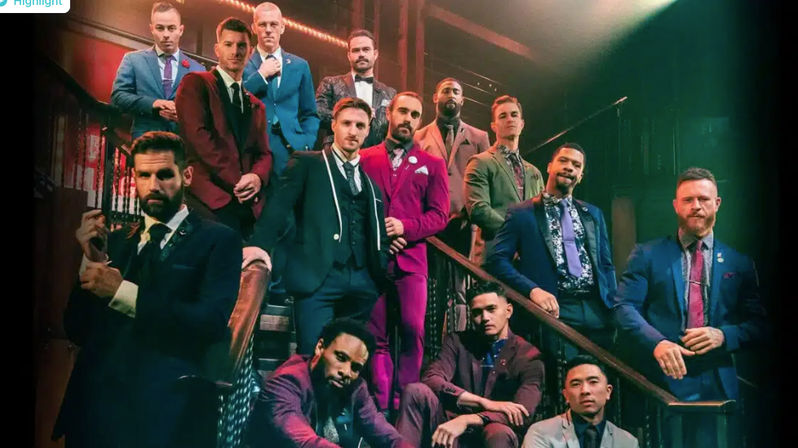 Group of dapper men in colorful suits and ties posing on a dramatic indoor staircase with theatrical lighting — stylish men's formalwear group portrait