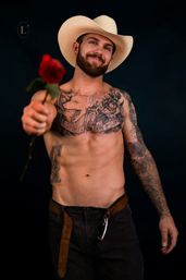 Smiling shirtless, heavily tattooed man in a white cowboy hat offering a red rose in a studio portrait with a dark background