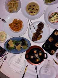 Overhead restaurant table shot of a fine-dining dinner spread on a white tablecloth, featuring tomato rigatoni, creamy soup, grilled steak medallions, green salad with parmesan crisps, bread, bite-sized canapés, skillet fritters, and printed menus.