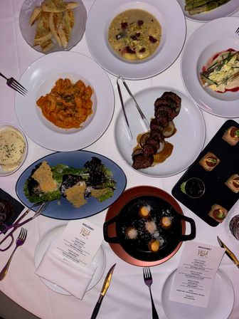 Overhead restaurant table shot of a fine-dining dinner spread on a white tablecloth, featuring tomato rigatoni, creamy soup, grilled steak medallions, green salad with parmesan crisps, bread, bite-sized canapés, skillet fritters, and printed menus.