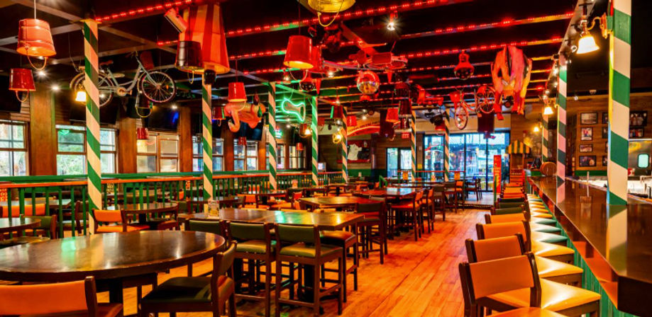 Vibrant empty pub-style restaurant interior with wooden floors, long bar and stools, high tables, and quirky hanging decor (bicycles, lamps, umbrellas) under red neon lighting.