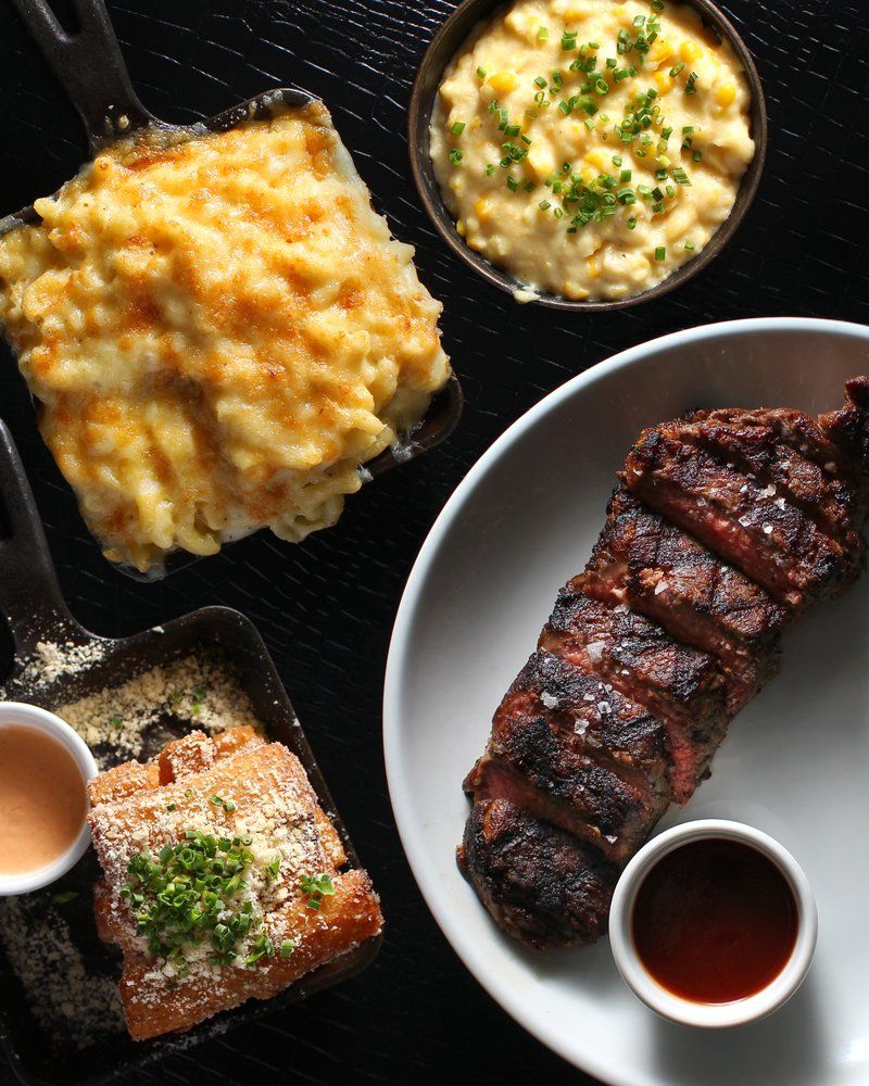 Top-down view of a sliced grilled steak with charred crust and a ramekin of sauce, plated beside cast-iron sides: bubbling golden mac and cheese, creamy corn with chives, and crispy parmesan-topped fried bread with dipping sauce – hearty comfort food on a black textured surface.