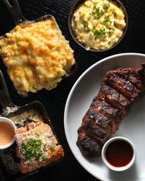 Top-down view of a sliced grilled steak with charred crust and a ramekin of sauce, plated beside cast-iron sides: bubbling golden mac and cheese, creamy corn with chives, and crispy parmesan-topped fried bread with dipping sauce – hearty comfort food on a black textured surface.