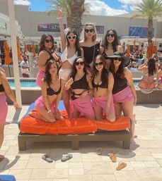 Eight women posing on an orange-cushioned cabana lounger at a sunny resort pool party, wearing bikinis, pink sarongs, sunglasses and sun hats, with palm trees, orange umbrellas and swimmers in the background.