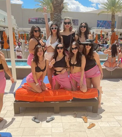 Eight women posing on an orange-cushioned cabana lounger at a sunny resort pool party, wearing bikinis, pink sarongs, sunglasses and sun hats, with palm trees, orange umbrellas and swimmers in the background.