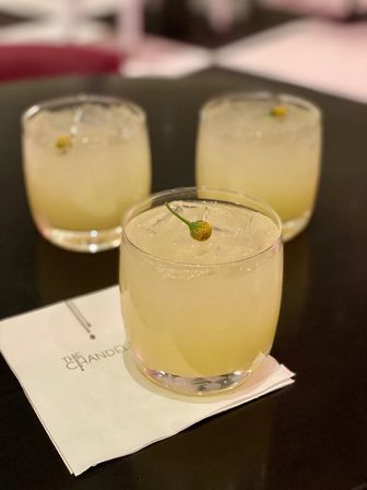 Three pale-yellow citrus cocktails in short rocks glasses with ice and tiny yellow flower-bud garnishes, arranged on a dark table with one glass on a white napkin.