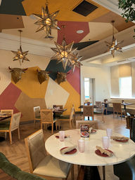 Restaurant dining room with hanging star-shaped pendant lights, decorative bull-head wall mounts on a colorful geometric mural, round table set with napkins and pink glasses, wooden chairs and warm wood floors.