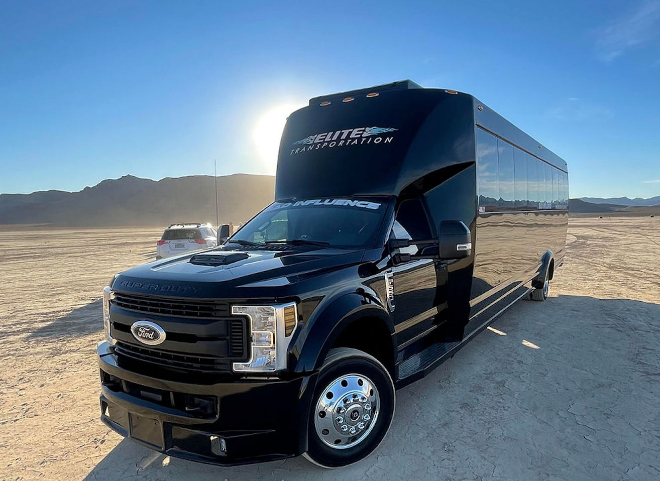 Glossy black coach-style shuttle parked on a sunlit desert playa with low mountains on the horizon, reflecting the blue sky and golden light.
