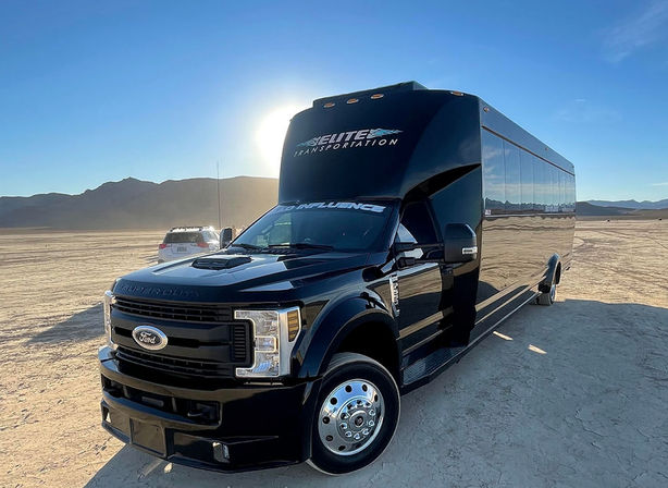 Glossy black coach-style shuttle parked on a sunlit desert playa with low mountains on the horizon, reflecting the blue sky and golden light.