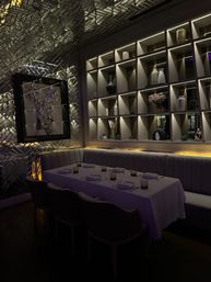Moody, dimly lit upscale restaurant private dining booth with white-clothed table set for six, tufted banquette seating, backlit decorative shelving of vases and a metallic textured wall featuring a framed modern portrait.