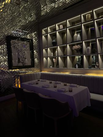 Moody, dimly lit upscale restaurant private dining booth with white-clothed table set for six, tufted banquette seating, backlit decorative shelving of vases and a metallic textured wall featuring a framed modern portrait.