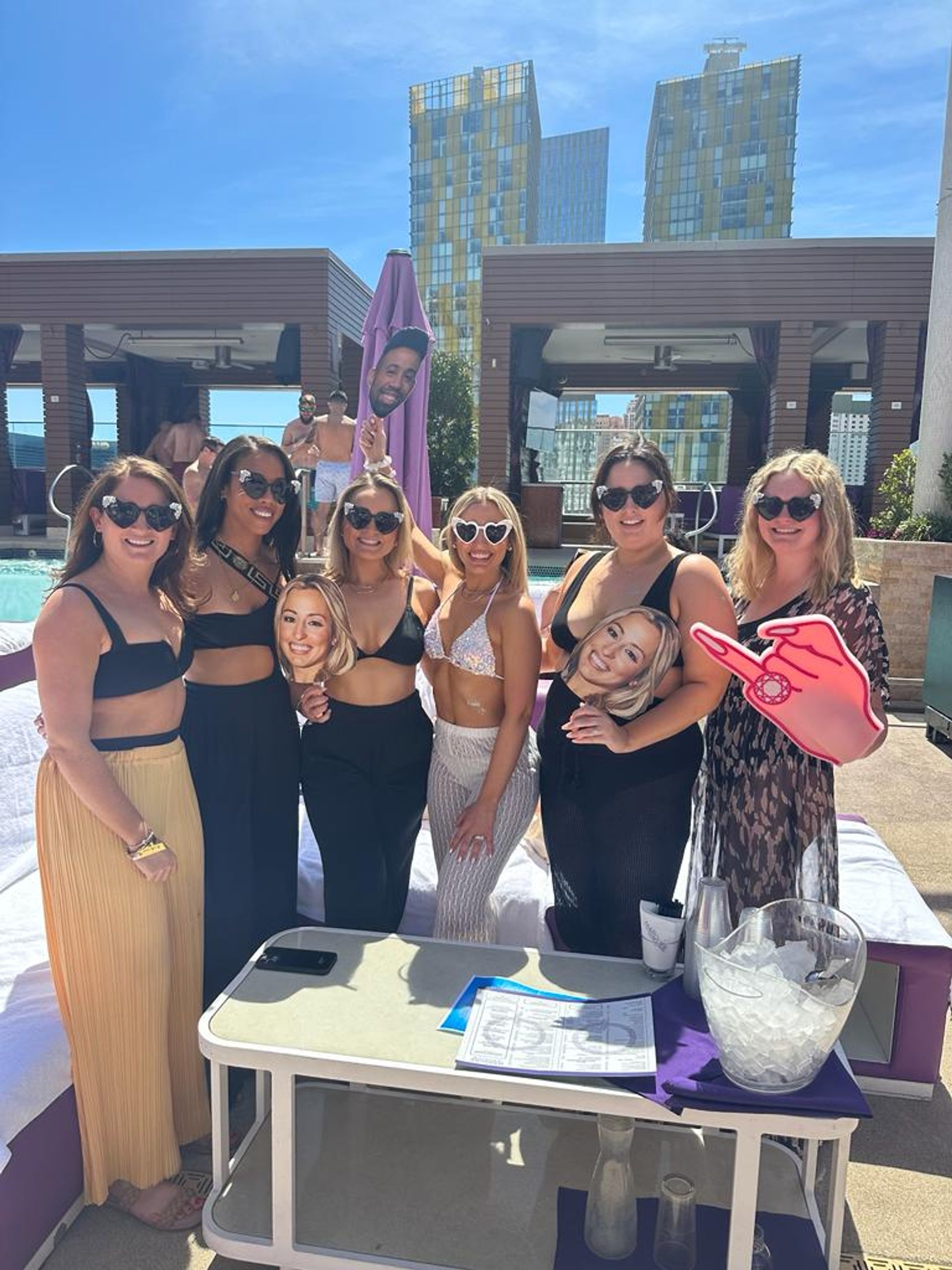 Seven friends in swimwear and heart-shaped sunglasses posing at a sunny rooftop pool and cabana area with downtown glass-tower skyline, holding oversized face cutouts and a foam hand prop.